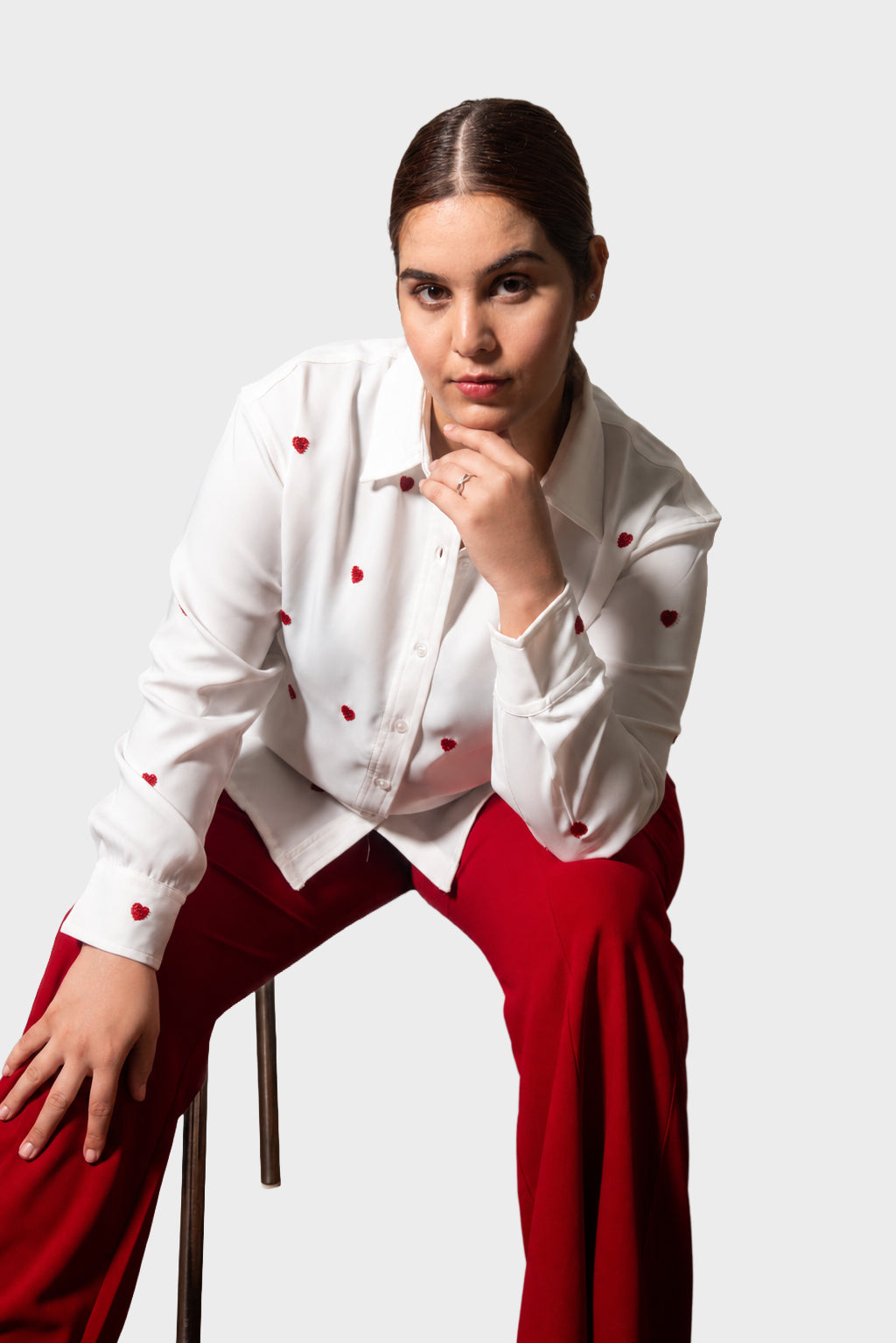 A woman posing in a white long-sleeve button-up shirt with small red embroidered details and bright red pants, seated on a stool against a light gray studio background.