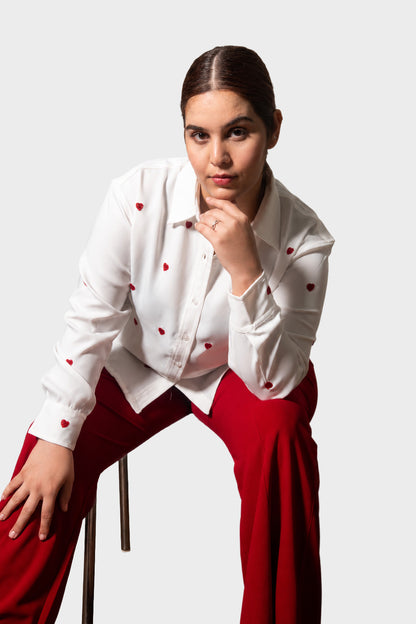 A woman posing in a white long-sleeve button-up shirt with small red embroidered details and bright red pants, seated on a stool against a light gray studio background.