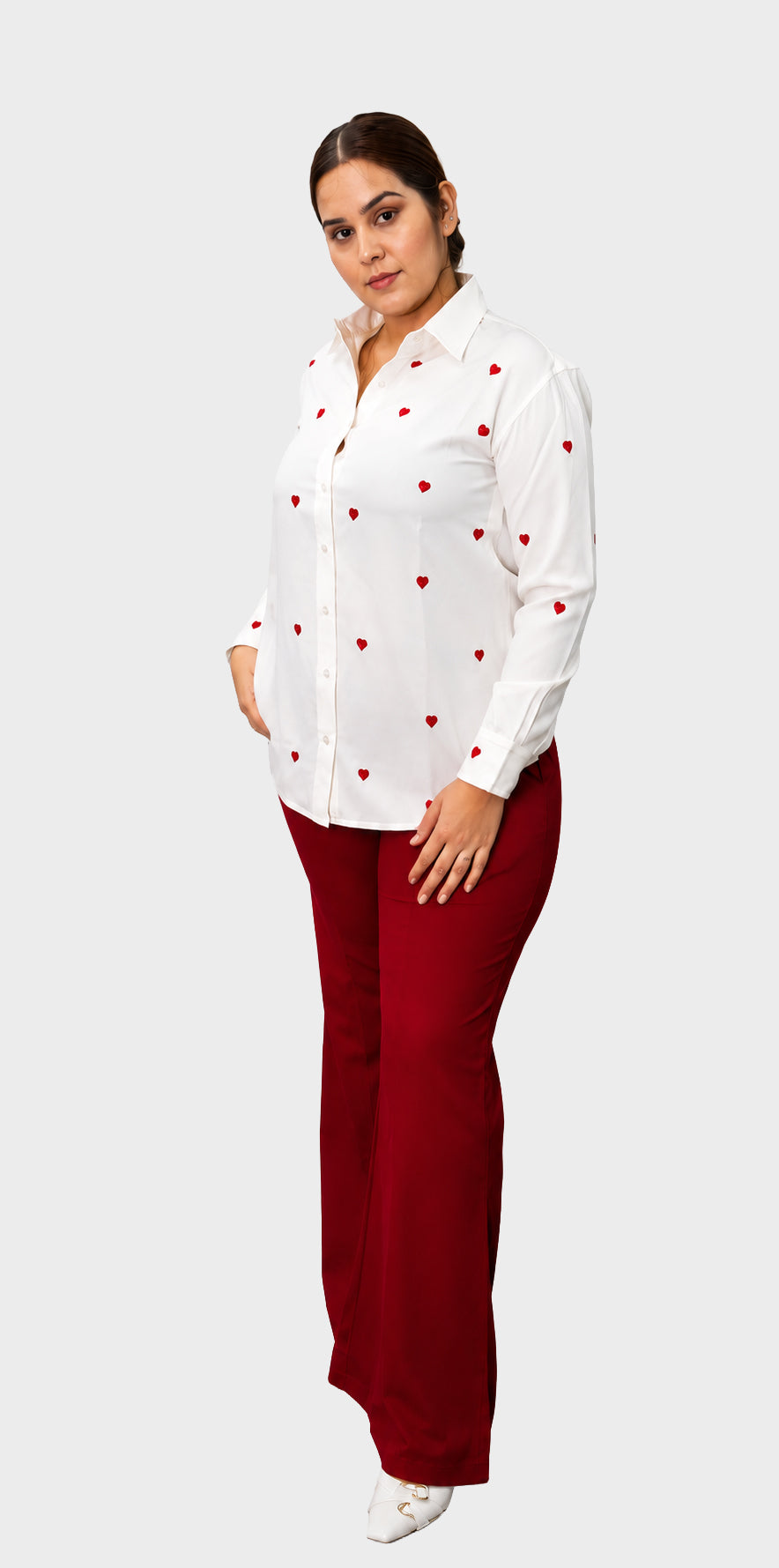 A woman modeling a white long-sleeve button-up shirt with small red heart patterns and dark red pants, standing against a light gray background.