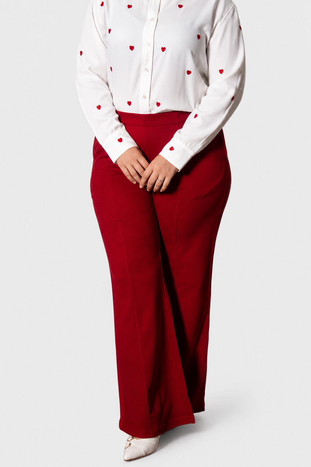 A model wearing a white long-sleeve button-up shirt with small red heart prints and high-waisted, wide-leg red trousers, paired with white pointed-toe heels, against a light gray studio background.