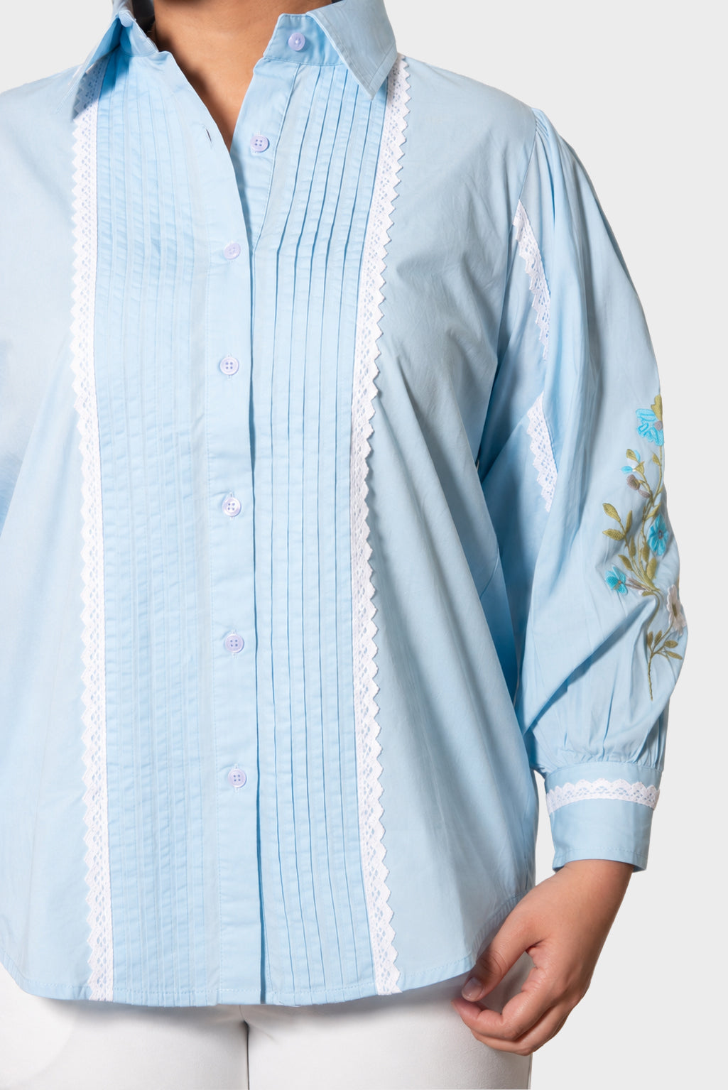 A light blue button-up shirt with white lace trim down the front and on the sleeve, featuring vertical pleats and floral embroidery on the cuff, shown on a model.