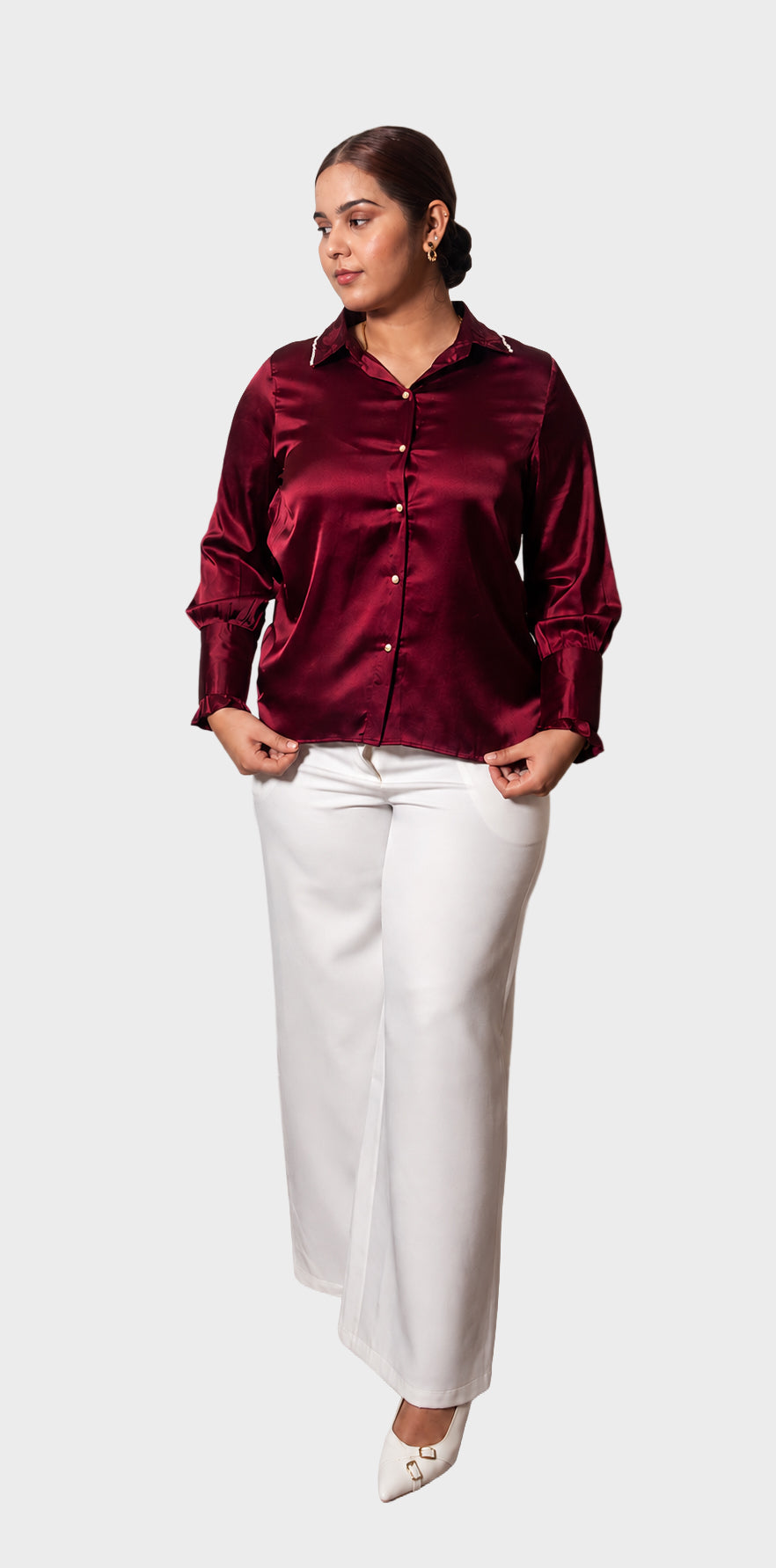 A woman modeling a long-sleeve satin button-up blouse in deep burgundy paired with white wide-leg pants and white pointed heels, set against a solid light gray background.