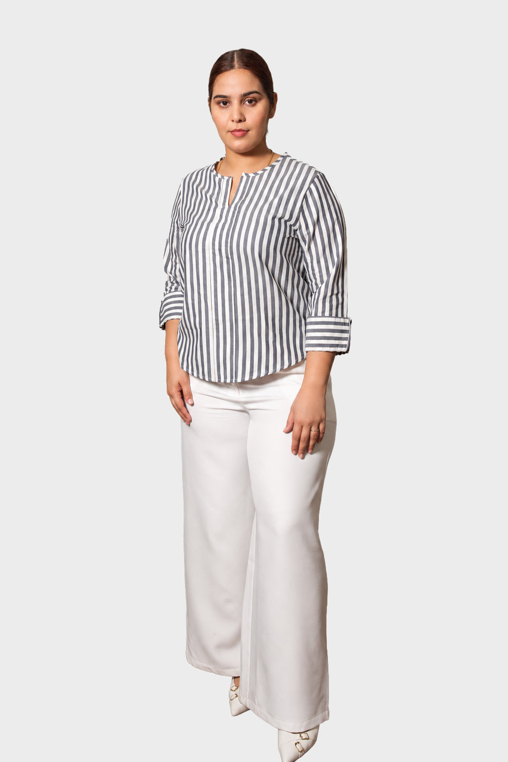 A woman modeling a black and white vertically striped long-sleeve blouse with a split neckline, paired with white wide-leg pants, against a solid light gray background.