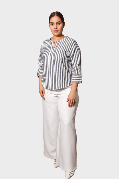 A woman modeling a black and white vertically striped long-sleeve blouse with a split neckline, paired with white wide-leg pants, against a solid light gray background.