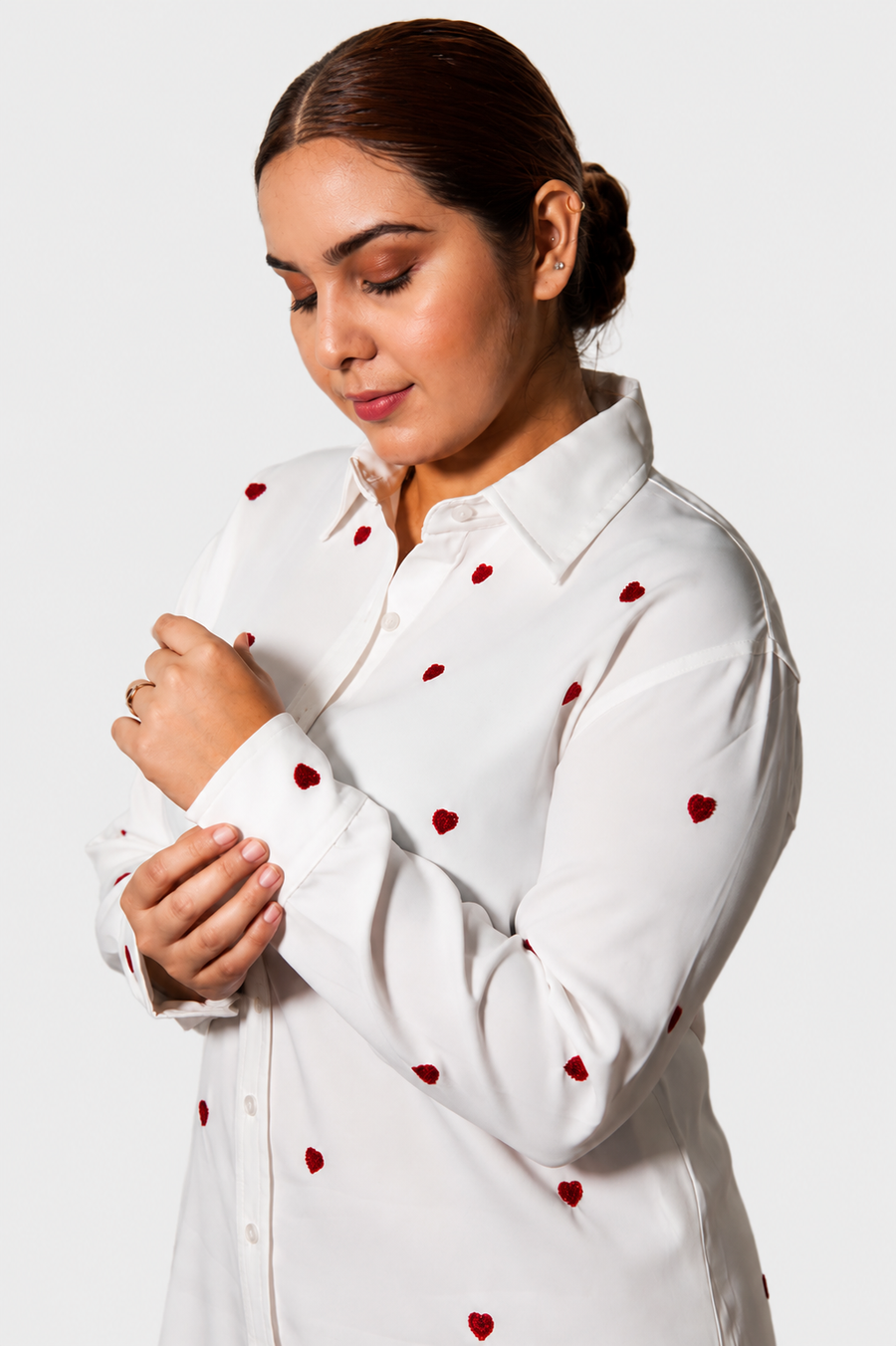 A woman wearing a white long-sleeve button-up shirt with small red heart embroidery, shown from the chest up against a light gray studio background.