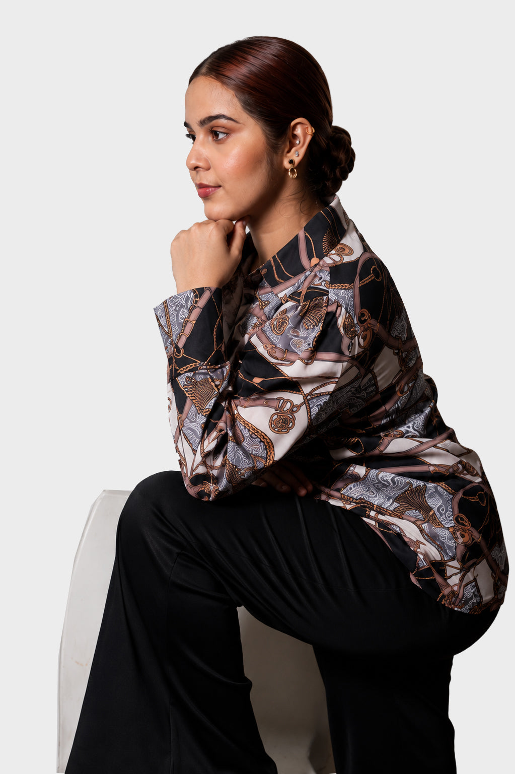 A woman seated on a white chair wearing a long-sleeve satin blouse with an abstract brown, beige, and black pattern, paired with black pants, against a light gray studio background.