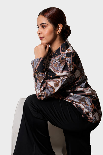A woman seated on a white chair wearing a long-sleeve satin blouse with an abstract brown, beige, and black pattern, paired with black pants, against a light gray studio background.